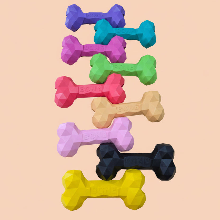 Nine colorful bone dog toys arranged in row.. The toys come in purple, teal, pink, green, coral, peach, light pink, black, and yellow, each with a faceted geometric texture and the word 'BONE' embossed on one end.