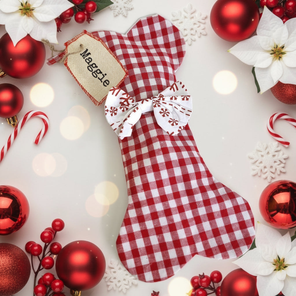 Red and white checkered dog Christmas bone shaped stocking with 'Maggie' tag surrounded by festive decorations on a light background.